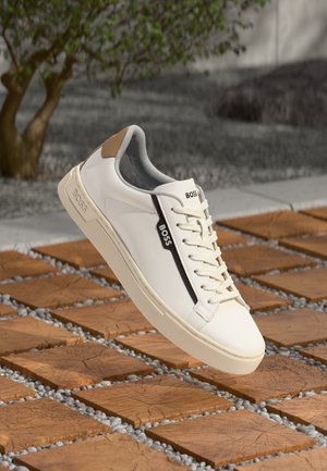 White leather sneaker with beige accents, a black stripe, and textured sole. Features round toe and classic lace-up design.