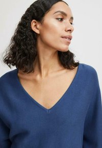 Suéter de punto azul con un escote en V profundo, que muestra una textura del tejido y un ajuste ligeramente oversize. El modelo tiene el cabello ondulado, presentado de perfil.
