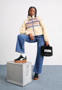 Veste zippée en peluche crème avec des rayures multicolores, jean en denim bleu évasé, baskets noires à semelles en gomme et sac en peluche noir avec logo blanc.