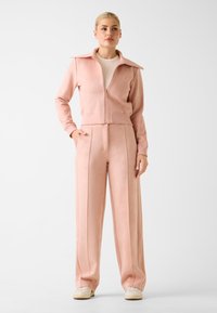 Tuta rosa chiaro composta da giacca con zip, colletto ampio e maniche lunghe, abbinata a pantaloni a vita alta e gamba larga con pieghe frontali.