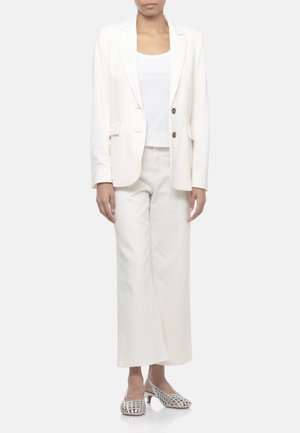 Donna che indossa un blazer color avorio, top bianco, pantaloni a gamba larga e scarpe slingback bianche con tacco basso e tessuto aperto, in piedi davanti a uno sfondo semplice.