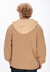 Tan fleecejakke med en tekstureret overflade og en kontrasterende beige hætte. Har en afslappet pasform og design med sænket skulder.
