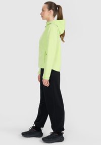 Femme de profil, portant une veste à capuche vert clair, un pantalon noir et des baskets noires sur un fond uni gris clair.