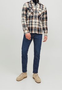 Camicia di flanella con un motivo a quadri nei colori crema, blu navy e ruggine; due taschini sul petto; abbinata a jeans blu scuro e scarpe beige.