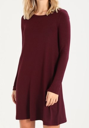 Kobieta ubrana w długą sukienkę o długości do kolan, w kolorze bordowym, z długim rękawem i okrągłym dekoltem, na jednolitym białym tle.