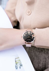 Runde schwarze Uhr mit Blumenmuster, roségoldenen Akzenten und einem Mesh-Armband. Zeigt den Markennamen an und hat dekorative Steine auf dem Zifferblatt.