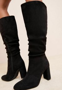 Stivali alti neri in suede con tacco a blocco e cerniere laterali indossati su gambe scoperte su sfondo beige.