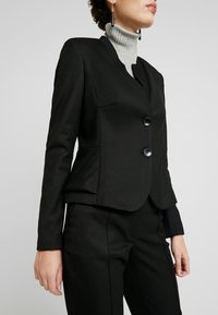 Zwarte aansluitende blazer van textuurstof, met een enkele knoopsluiting en twee voorzakken. Draagt over een lichtgrijze geribbelde turtleneck.