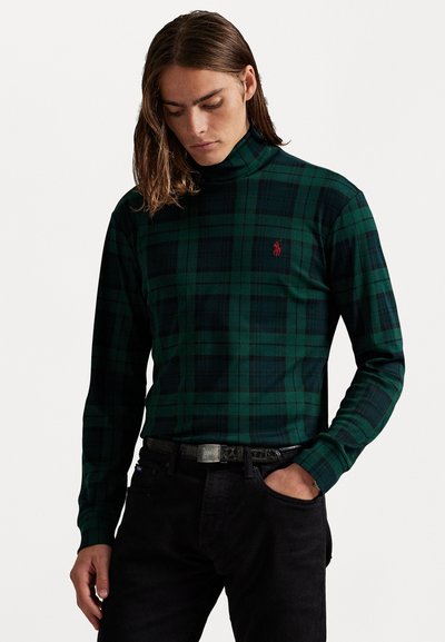 Groen en zwart geruit coltrui met lange mouwen en een klein rood logo op de borst, gecombineerd met zwarte jeans en een riem.