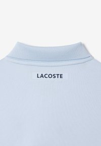 Col de chemise bleu clair avec une étiquette blanche cousue à l'intérieur, portant le nom de la marque "LACOSTE" en lettres majuscules noires.