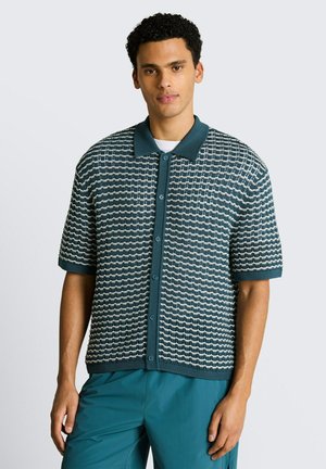 Junger Mann trägt ein türkis-weiß gemustertes kurzärmeliges Hemd und türkisfarbene Hose vor hellem einfarbigem Hintergrund.