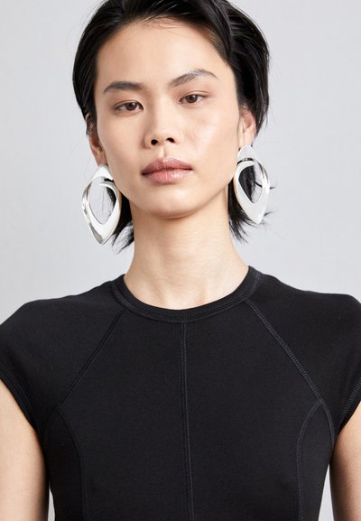 De grandes boucles d'oreilles en argent abstraites et un haut ajusté à manches courtes de couleur noire avec une finition texturée. Le design présente des coutures distinctives.