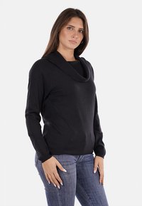 Maglione nero a maniche lunghe con scollo a cowl e una texture liscia, abbinato a jeans blu. Design semplice e aderente, senza pattern.