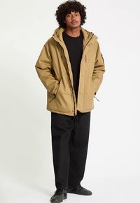 Tan zip-up jacket with a hood, features a drawstring hem, and side pockets. Worn over a black shirt and paired with black pants and shoes.