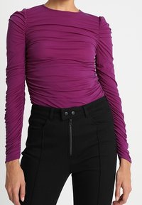 Lila Langarmshirt mit gerafften Details am Oberteil und an den Schultern, kombiniert mit hochtaillierten schwarzen Hosen mit einem Reißverschluss vorne.