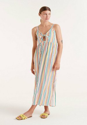 Vestido maxi a rayas en blanco, con rayas verticales de colores del arcoíris, un detalle de lazo en el pecho y tirantes de espagueti ajustables. Sandalias planas amarillas.