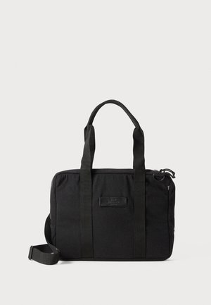 Svart tygväska med dubbla handtag upptill, justerbar axelrem och dragkedjestängning. Har en framtill placerad etikett och en minimalistisk design.