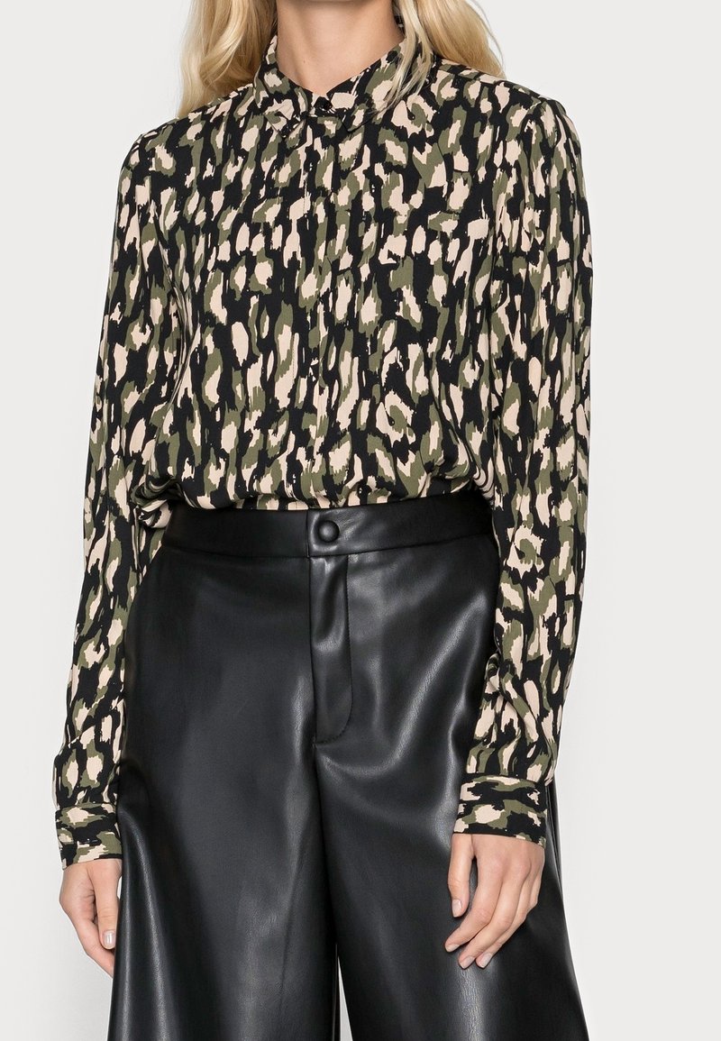 Blouse à manches longues avec un design léopard vert, noir et beige, dotée d'un col et d'un tissu doux, associée à un pantalon en cuir noir taille haute.
