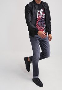Chaqueta bomber de cuero negro, camiseta gráfica con patrones rojos y negros, jeans grises y zapatillas negras en una pose casual.