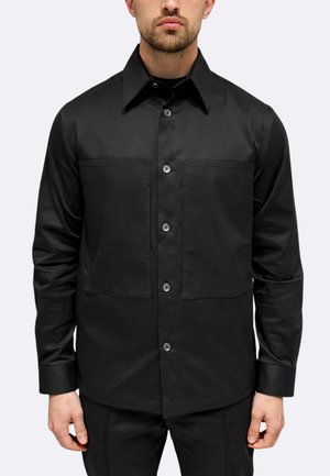 Uomo con camicia nera abbottonata con due grandi tasche frontali, in piedi davanti a uno sfondo chiaro e uniforme.