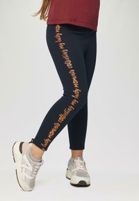 Mariene legging met "gelukkige momenten verzamelen" in oranje schrift langs de zijkant, gecombineerd met lichtgekleurde sportschoenen. Glad materiaal, nauwsluitende pasvorm.