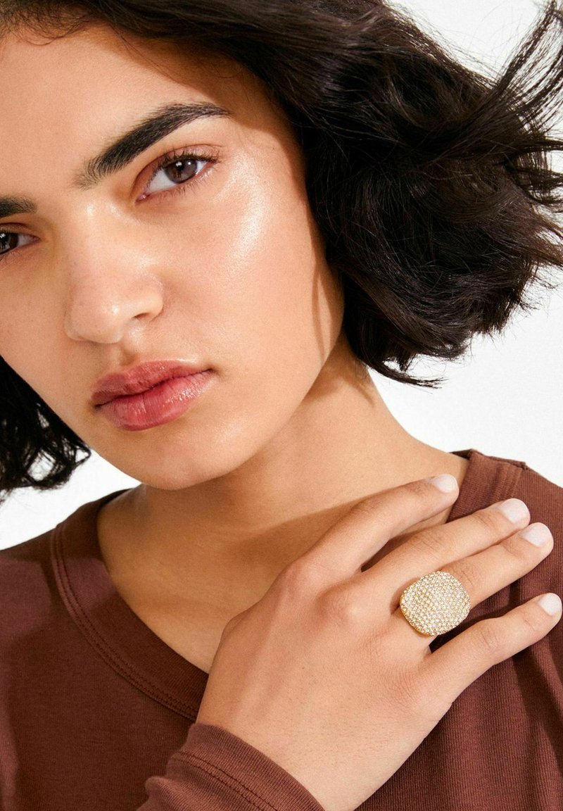 Anillo de tono dorado con un diseño texturizado y múltiples pequeñas gemas, en forma de cuadrado redondeado, llevado en una mano sobre una tela marrón.