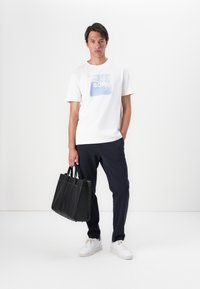 Witte katoenen t-shirt met blauwe grafische print, donkerblauwe broek en zwarte leren handtas. Model draagt witte sneakers en staat tegen een effen achtergrond.