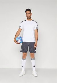 Valge sportliku t-särgiga, millel on mustad õlastriibid, hallid lühikesed püksid ja valged sokid mustade triipudega. Hoidmas mitmevärvilist jalgpalli.