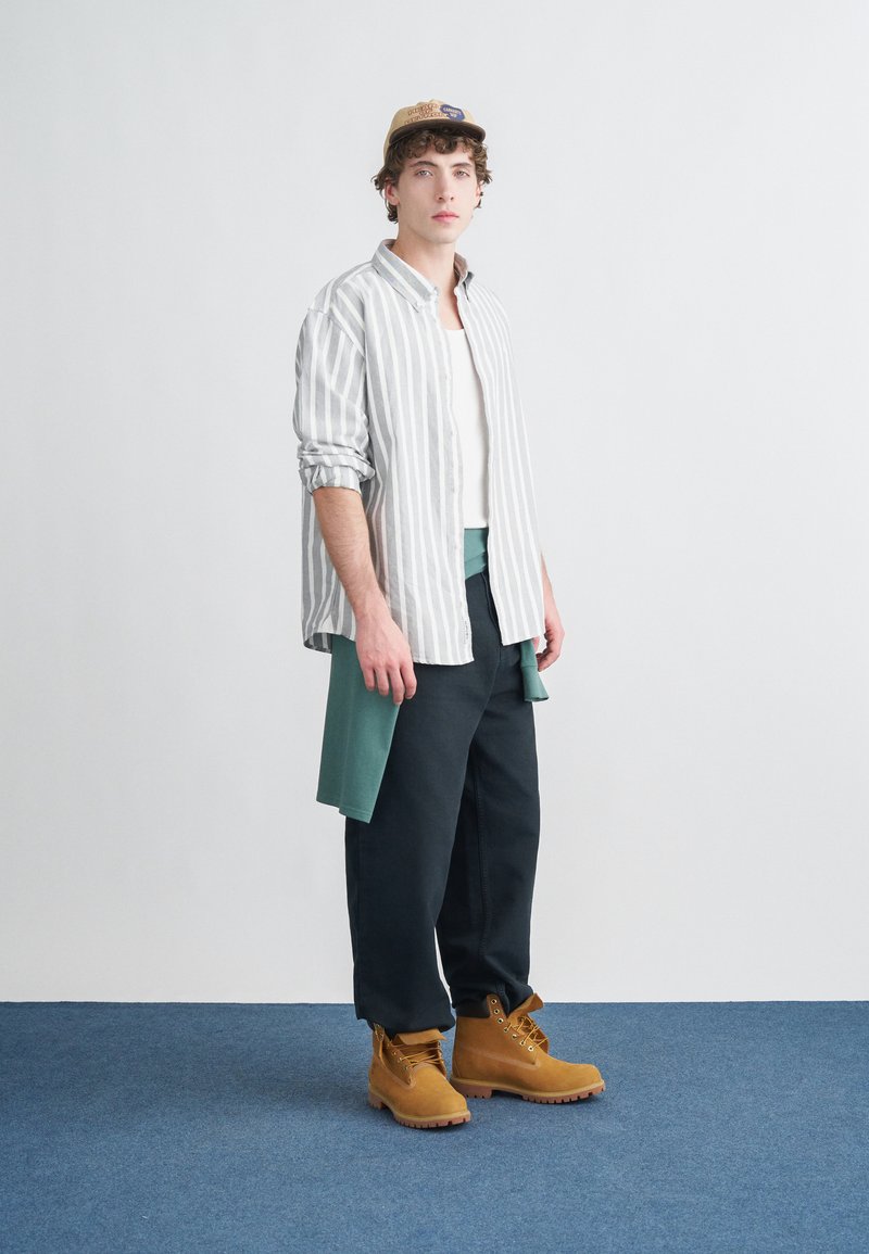 Maglietta a maniche lunghe a righe grigio chiaro e bianco sopra a una canotta bianca, abbinata a pantaloni scuri e stivali marroni. Un berretto beige completa il look.