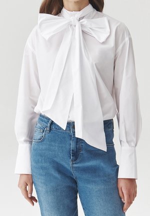 Frau trägt eine weiße langärmelige Bluse mit großer Schleife am Hals und blaue hoch taillierte Jeans vor einfachem Hintergrund.