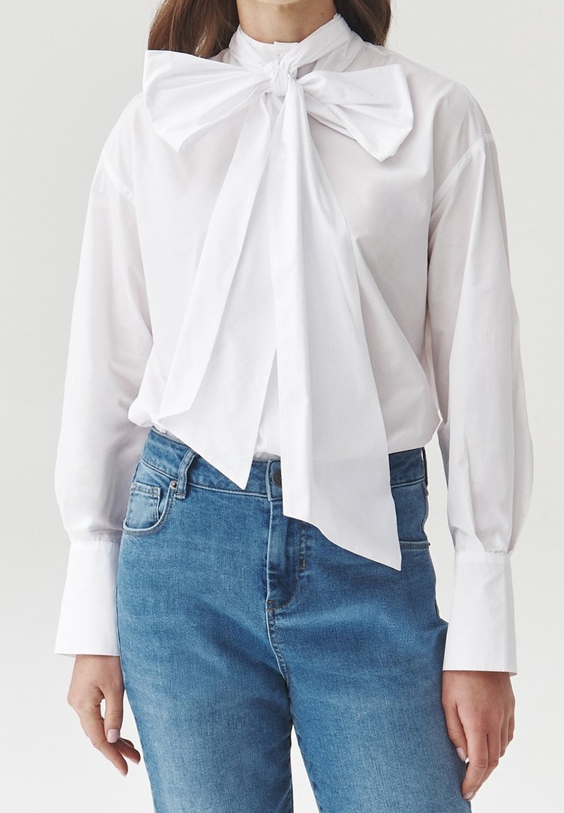 Femme portant un chemisier blanc à manches longues avec un grand nœud au cou et un jean bleu taille haute sur un fond uni.