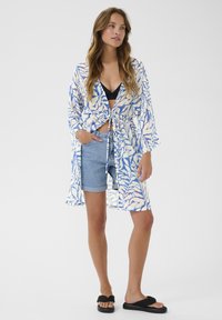 Kimono ligero y semitransparente con un patrón de hojas en azul y blanco, atado en la cintura. Combinado con pantalones cortos de mezclilla y sandalias negras tipo slide.