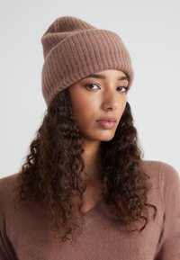 Beanie a coste marrone in materiale morbido, con un design slouchy e orlo arrotolato. Il modello indossa un maglione coordinato, che mostra un dettaglio testurizzato.
