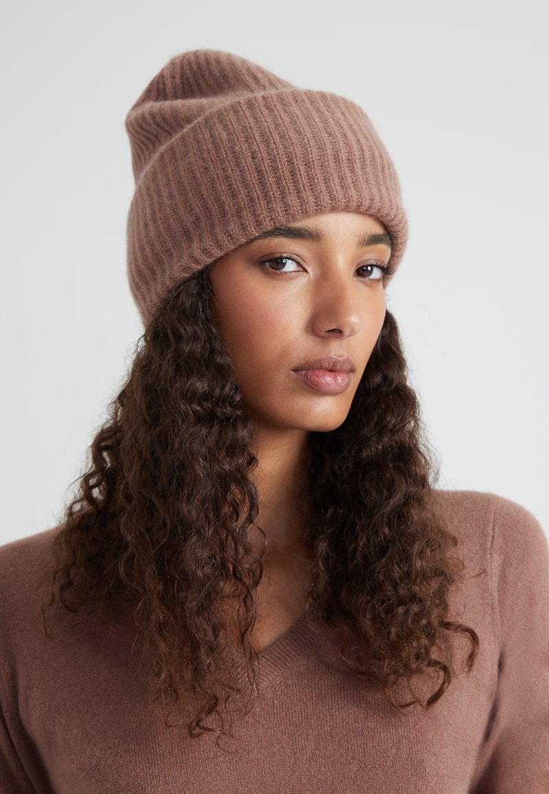 Beanie a coste marrone in materiale morbido, con un design slouchy e orlo arrotolato. Il modello indossa un maglione coordinato, che mostra un dettaglio testurizzato.