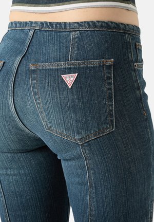 Tył osoby noszącej niebieskie jeansy Guess z denimu, z naszywką z logo trójkąta na prawym tylnym kieszeni oraz paskowanym zakończeniem koszuli.