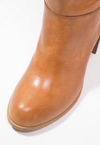 Botte de cheville en cuir tan avec une finition lisse, bout rond et un petit talon carré. Présente des coutures discrètes le long des coutures.