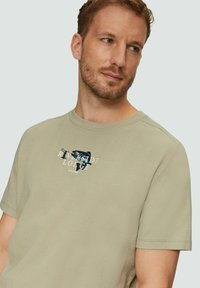 Lichtgroene T-shirt met korte mouwen, gemaakt van gestructureerde stof, met een grafische print van "RIVERSIDE LODGE" en gestileerde afbeeldingen op de voorkant.