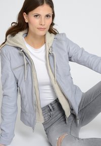 Lichtgrijze leren jack met beige capuchonvoering, rits aan de voorkant en zijzakken. Kenmerkt zich door gequilte details op de schouders.