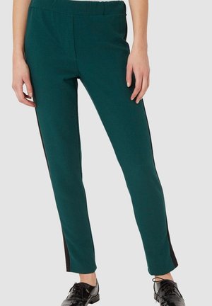 Piernas con pantalones verdes ajustados con rayas laterales negras y zapatos negros brillantes con cordones sobre un fondo claro y liso.