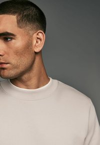 Sudadera gris claro con un cuello redondo acanalado, que presenta una textura de tela suave. Primer plano de la cara de un hombre, mostrando cabello corto y detalles faciales.