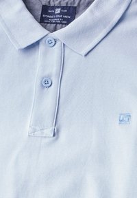 Ljusblå polo med texturerat tyg. Har en klassisk krage, treknappslå och en liten logotyp på bröstet.