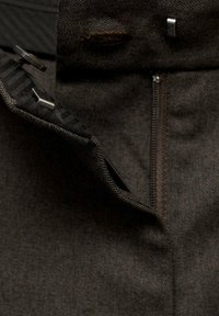 Pantalon en tissu foncé avec une fermeture éclair partiellement ouverte et une ceinture déboutonnée montrant des fermetures en métal à crochets et barres.