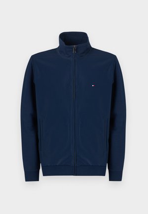 Námornícka bunda na zips s vysokým golierom, vyrobená z hladkej látky, má bočné vrecká a malú logo v červenej, bielej a modrej farbe na hrudi.