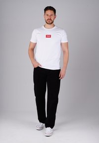 Weißes kurzärmliges T-Shirt mit einem kleinen roten Grafiklabel auf der Brust, kombiniert mit schwarzen Hosen und weißen Sneakern. Schlichter und lässiger Look.