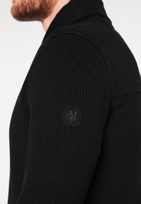 Schwarzer Strickpullover mit strukturierter Oberfläche und geripptem Muster, mit einem runden, geprägten Logo auf dem linken Ärmel.