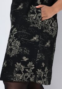 Schwarzes Kleid aus strukturiertem Stoff mit goldenen Blumen- und Vogelmustern, seitlichen Taschen, knielanger Schnitt und subtiler Schlitze am Saum.