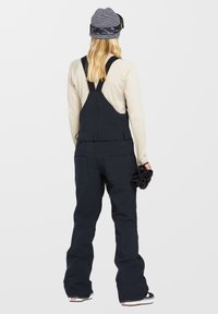 Navy Snowboardoverall mit verstellbaren Trägern, Reißverschlusstaschen und lockerer Passform. Kombiniert mit einem leichten Langarmshirt und einer gemusterten Mütze.