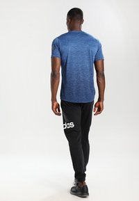 Blå, kortärmad tränings-T-shirt i mjukt tyg, kombinerad med svarta joggerbyxor som har en stor vit logotyp på sidan.