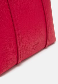 Gros plan sur un sac en cuir rouge texturé avec couture verticale et logo embossé subtil près du bord inférieur sur un fond blanc.