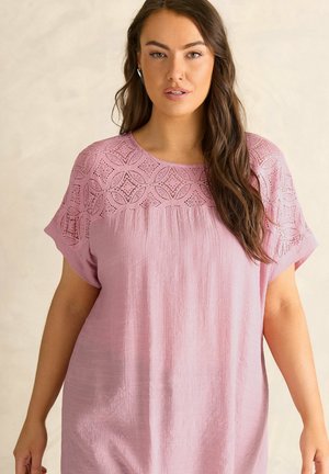 Kvinde med langt brunt hår iført en løs lyserød bluse med blonder på den øverste del af brystet og ærmer mod en beige baggrund.